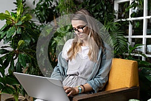 Woman gardener sitting on chair in green house, working on laptop surrounded by plants, remote work, distance job