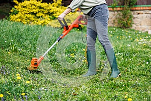 Woman gardener using electric string trimmer to trim grass in garden