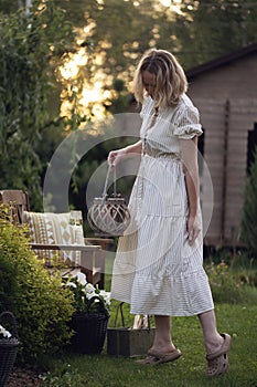woman gardener in white romantic dress relaxing in summer garden, holding lantern.
