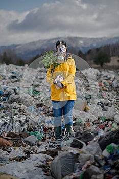 Woman with gas mask holding green plant on landfill, environmental concept.