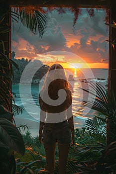 A woman stands by the ocean at sunset, gazing at the colorful sky and water