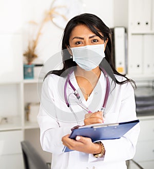 Woman general practitioner in mask filling clipboard with medical records