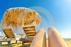 Woman girl legs sunbathing on tropical sandy beach with blurred straw sunshade umbrella and wooden sun lounger