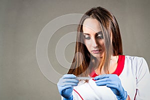 Woman or girl nurse or doctor in medical dressing gown with sterile gloves on her hands looks at the thermometer in her hands on