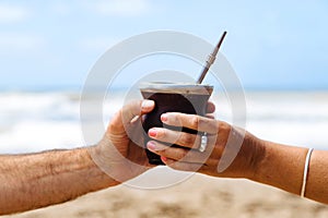 Woman giving to a man a Yerba Mate hot drink in the beach.