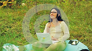 A woman with glasses on the edge of a forest with a pencil in her hand and a laptop thinks over the project.