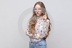 Woman in glasses with long blond hair seriously pointing finger up and warning, looking at camera.