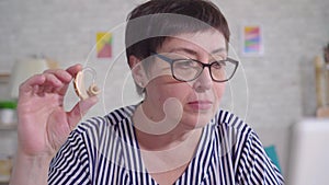 Woman with glasses sitting at a table in the living room holding a hearing aid uses a laptop close up