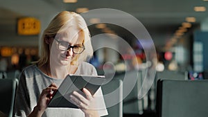 A woman with glasses and summer clothes is waiting for her flight at the terminal of the airport. Enjoys internet at the