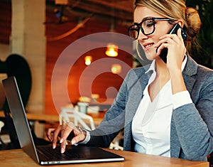 woman in glasses talking on mobile phone and using laptop with smile while sitting at her working place.