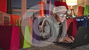 Woman with glasses wearing a santa claus hat is lying on the carpet and makes an online purchase using a credit card and