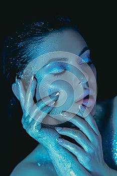 Woman with glittery makeup touches her face, bathed in an ethereal blue glow