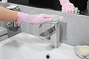 Woman in glove cleaning faucet of bathroom sink with paper towel, closeup