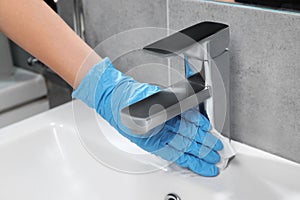 Woman in glove cleaning faucet of bathroom sink with paper towel, closeup