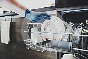 Woman gloved hand is loading dirty utensil to dishwasher machine at kitchen