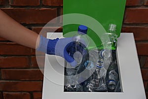 Woman in gloves putting used plastic bottle into trash bin near brick wall, closeup. Recycling problem