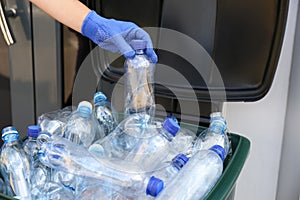 Woman in gloves putting used plastic bottle into trash bin outdoors, closeup. Recycling problem