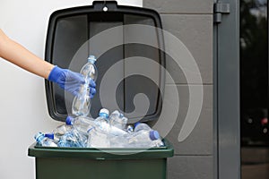 Woman in gloves putting used plastic bottle into trash bin outdoors, closeup. Recycling problem