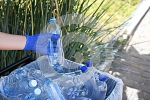 Woman in gloves putting used plastic bottle into trash bin outdoors, closeup. Recycling problem