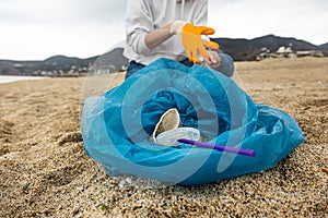 A woman in gloves with a special blue bag picks up garbage among the sand along the coast. The problem of environmental