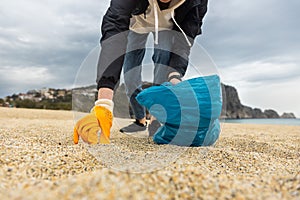 A woman in gloves with a special blue bag picks up garbage among the sand along the coast. The problem of environmental