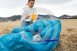 A woman in gloves with a special blue bag picks up garbage among the sand along the coast. The problem of environmental