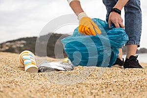 A woman in gloves with a special blue bag picks up garbage among the sand along the coast. The problem of environmental