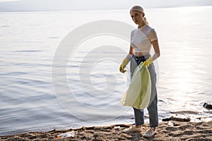 A woman in gloves stands on the shore of the lake and looks into the camera with annoyance. There is a lot of rubbish