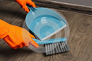 Woman in gloves sweeping wooden floor with plastic whisk broom and dustpan indoors, closeup