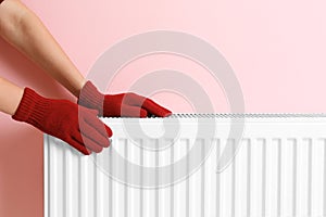 Woman in gloves warming hands on heating radiator near color wall