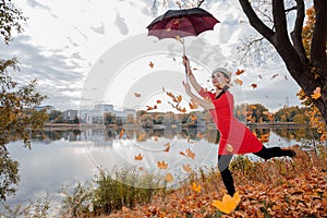 A woman in a gray beret, brunette with long hair, in a red dress holds an umbrella high above her head, maple leaves are