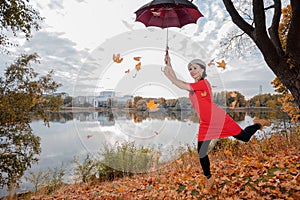 A woman in a gray beret, brunette with long hair, in a red dress holds an umbrella high above her head, maple leaves are