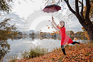 A woman in a gray beret, brunette with long hair, in a red dress holds an umbrella high above her head, maple leaves are