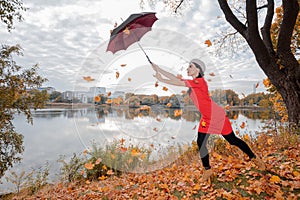 A woman in a gray beret, brunette with long hair, in a red dress holds an umbrella high above her head, maple leaves are