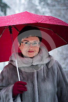 woman in a gray fur coat with a red umbrella stands on a white, snow-covered background.