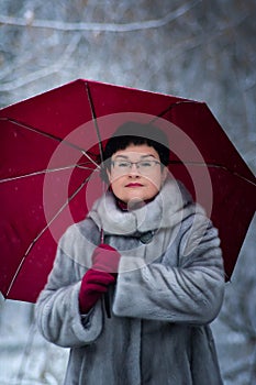 woman in a gray fur coat with a red umbrella stands on a white, snow-covered background.