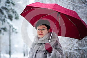 woman in a gray fur coat with a red umbrella stands on a white, snow-covered background.