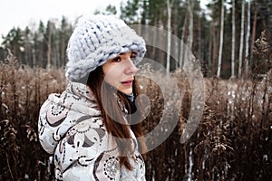 woman with gray knitted hat and warm coat posing in winter forest. Portrait of thoughtful girl outdoors on winter day