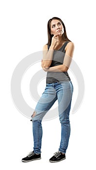 Woman in gray top and blue jeans standing with thoughtful expression on her face and with hand on her chin looking up