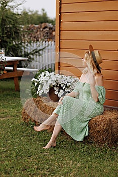 A woman in a green dress sits on hay bales near a wooden house