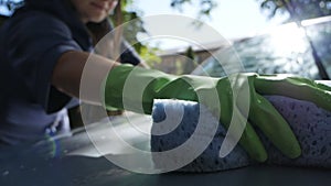 Woman in green gloves with a sponge wipes the car in the garden near the house in summer