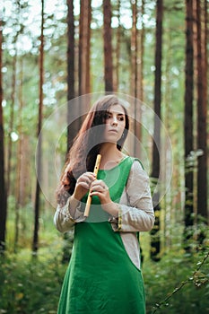 Woman in a green medieval dress plays the wooden flute while standing in a grove against the background of pines. Girl in a gloomy