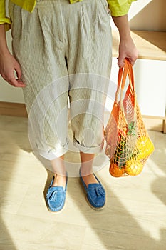 Woman in green shoes holding string grocery reusable mesh bag full of fresh fruits and vegetables