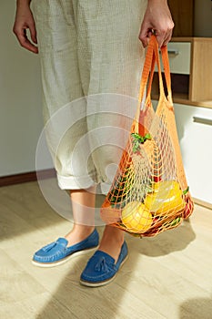 Woman in green shoes holding string grocery reusable mesh bag full of fresh fruits and vegetables