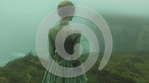 Woman in Green Vintage Dress Facing Mist-Enshrouded Cliffs