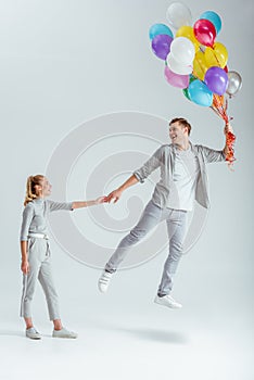 woman in grey clothing holding hand of happy man jumping in air with bundle of colorful balloons