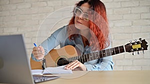 A woman with a guitar writes notes in a notebook. The girl composes a song