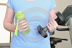 Woman in the gym holds a metal dumbbell and a water bottle in her hands