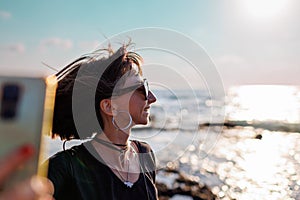 woman with hair blowing in the wind taking a selfie by the sea. Beautiful girl smiling while looking at camera outside -