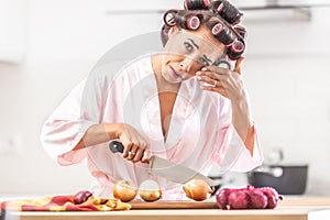 Woman in hair curlers and nighgown cries from onion cutting in the kitchen, wiping the tear from her eye